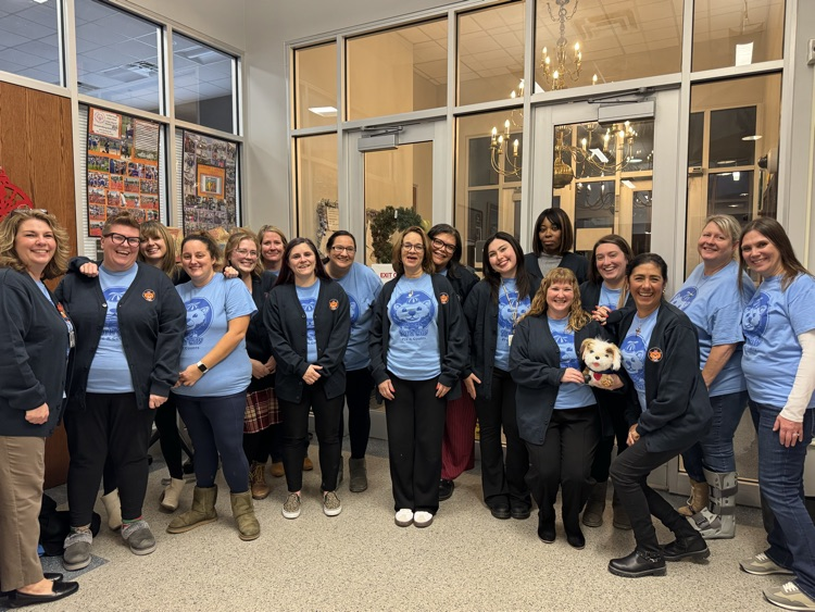 A group of fourteen district staff members, wearing matching blue shirts and dark jackets, are stand together in a lobby area. One woman is holding a small stuffed animal.