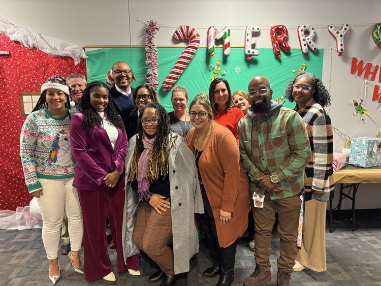 A group of twelve district staff members wearing festive holiday attire are standing in front of a green and red Christmas backdrop with decorations and presents.