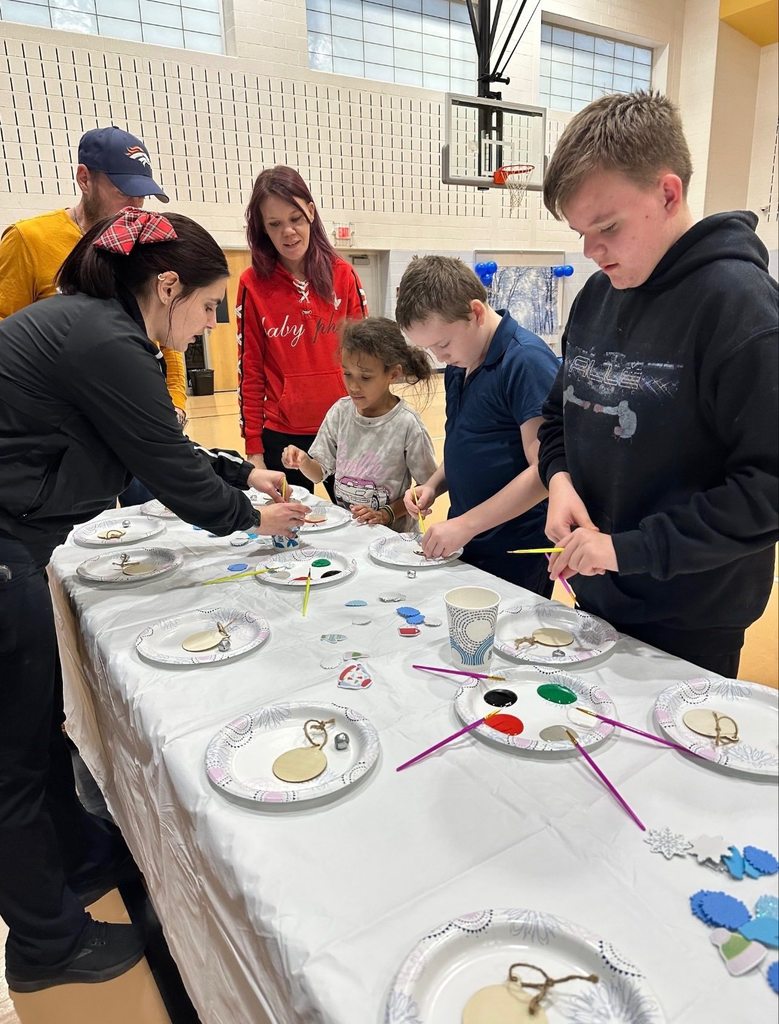 A group of children and adults are doing crafts at a table covered with art supplies in a school gym.