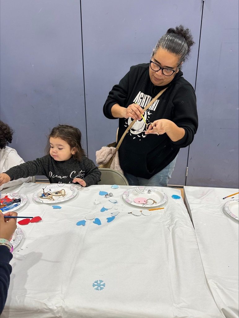 An adult and a young child are engaging in a craft activity at a table covered with a white cloth in a school gym. They are surrounded by paper hearts and plates of craft supplies.