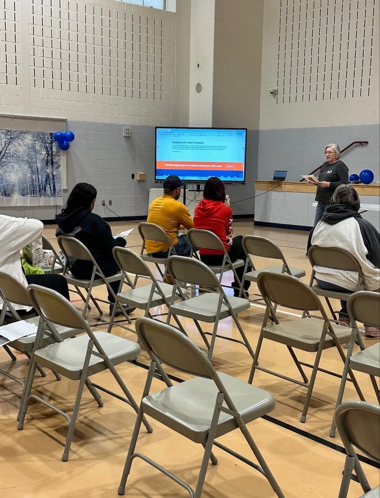 An adult woman is presenting in a school gym to a group of seated adults. A large screen displaying a webpage can be seen nearby.