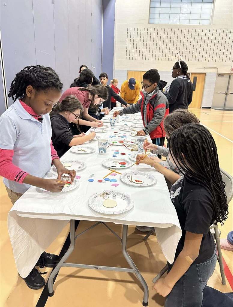 A group of children and are gathered around a table in a school gym, engaging in arts and crafts. Plates and art materials are spread out across the table.