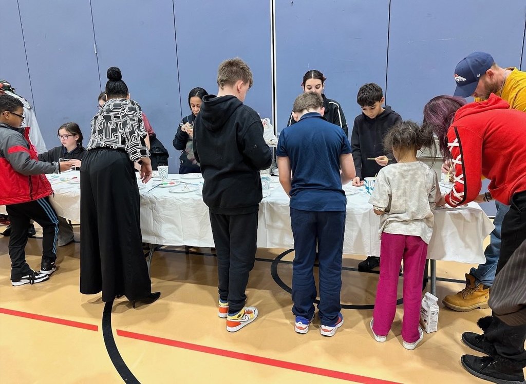 A group of people, including children and adults, are gathered around a table covered with a white cloth. They are engaging in a craft activity in a school gym.