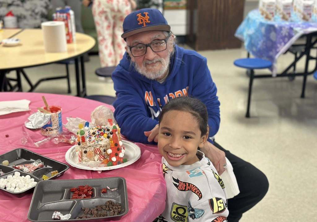 A man and a young boy are sitting around a pink tablecloth-covered table in a school cafeteria with a decorated gingerbread house nearby.