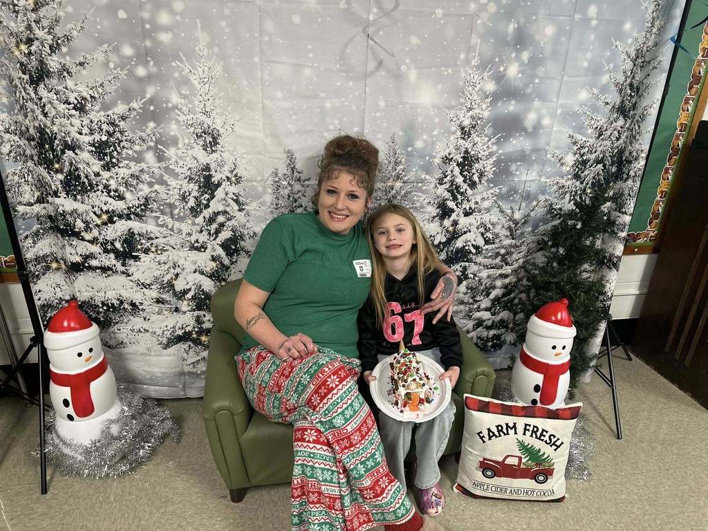 A woman and a young girl are happily posing with a decorated gingerbread house on a plate. They are sitting on a green chair in front of a snowy forest backdrop.