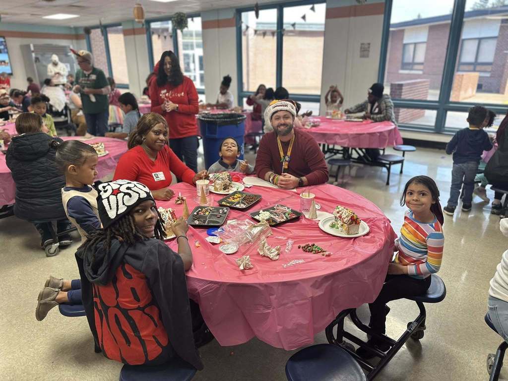 Children and adults, including a district staff member, are seated around tables covered in pink tablecloths, making crafts in a school cafeteria.