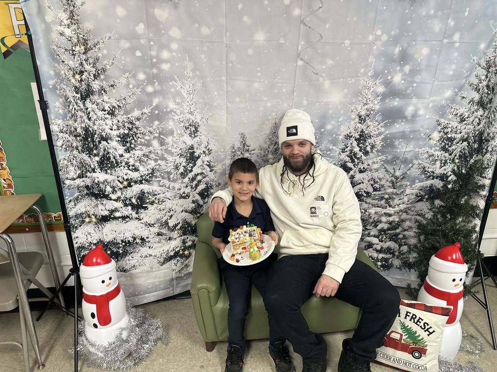 A man and a young boy are happily posing with a decorated gingerbread house on a plate. They are sitting on a green chair in front of a snowy forest backdrop.