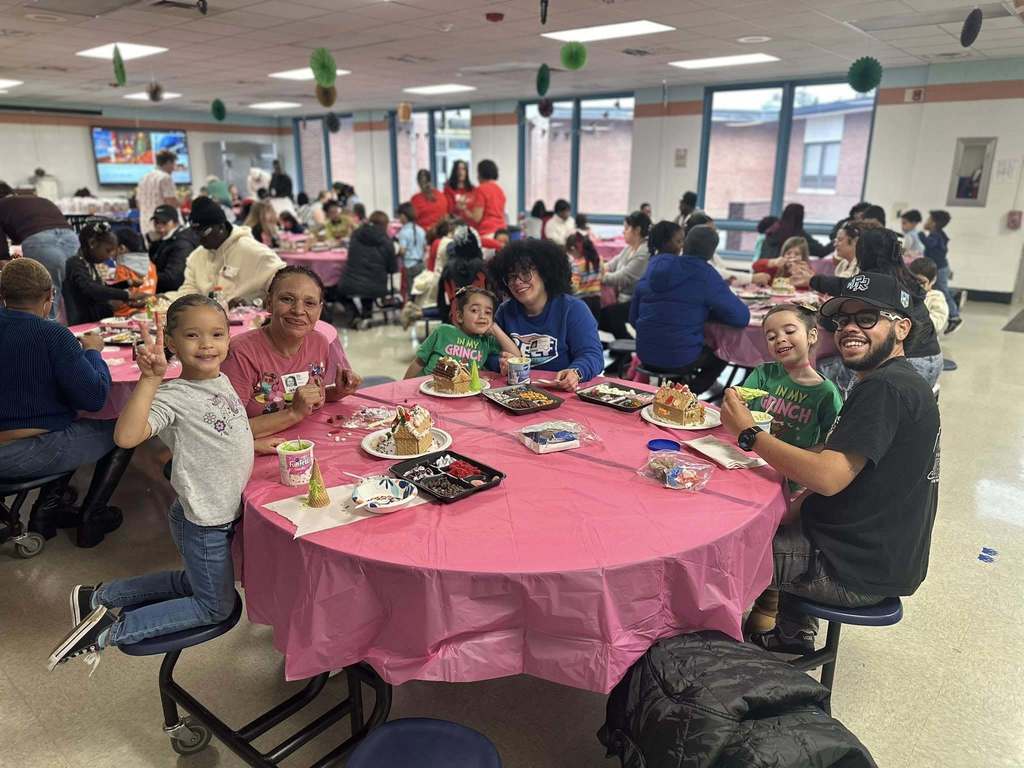 A lively gathering in a school cafeteria shows a family seated at a pink cloth-covered table, engaging in gingerbread house decorating. 