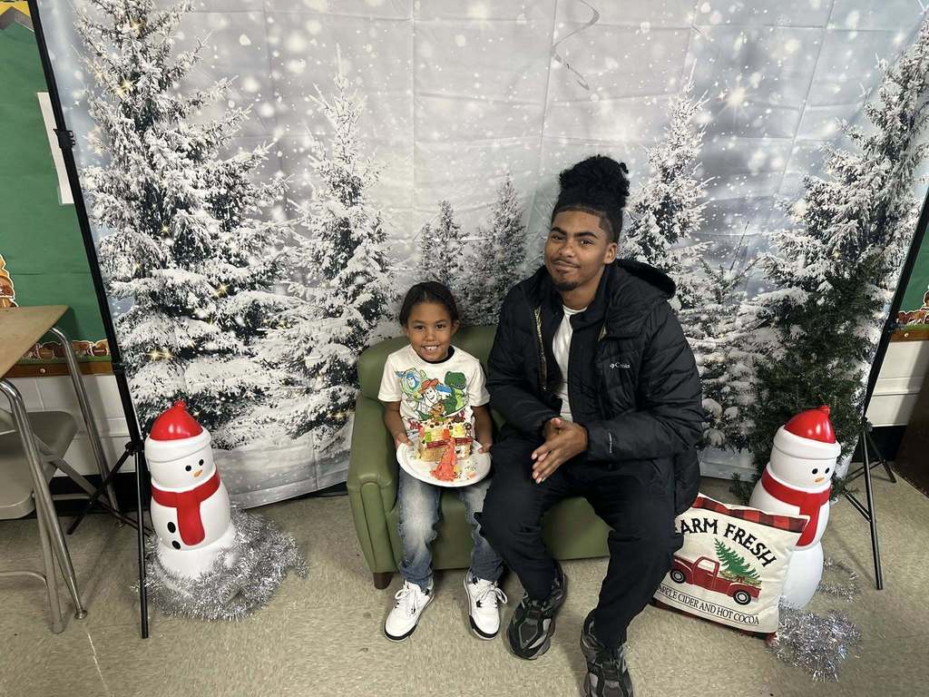 A man and a young boy are happily posing with a decorated gingerbread house on a plate. They are sitting on a green chair in front of a snowy forest backdrop.