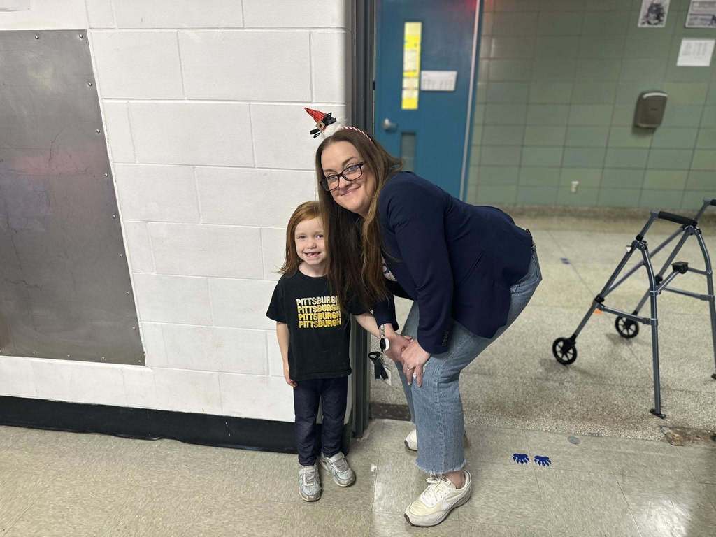 A district staff member wearing a festive headband is bending down next to a young girl in a school cafeteria. A walker can be seen in the background. 