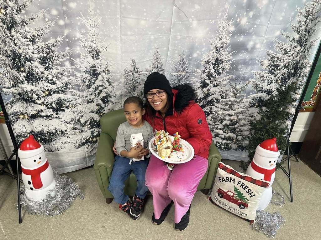 A woman and a young boy are happily posing with a decorated gingerbread house on a plate. They are sitting on a green chair in front of a snowy forest backdrop.