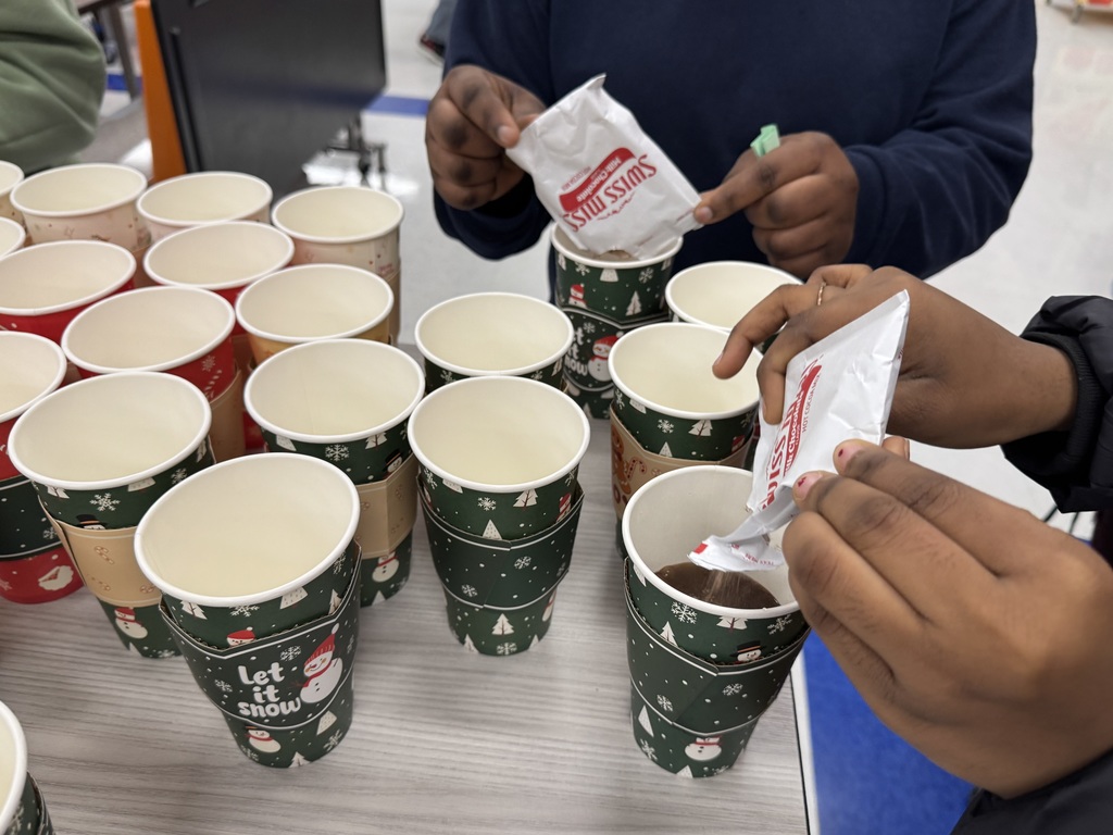 Two people can be seen dumping packets of hot cocoa mix into Christmas-themed paper cups. The table is filled with multiple paper cups that are decorated with snowflakes and snowmen.