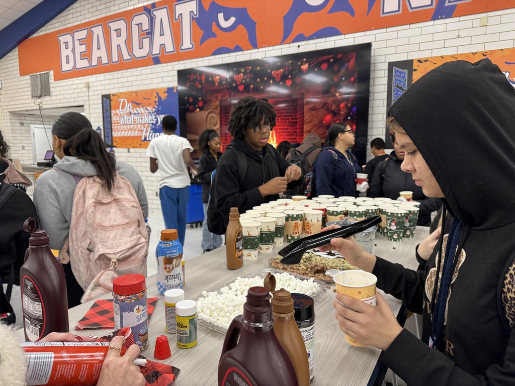 A group of students are gathered around a table filled with hot chocolate supplies and toppings in a school cafeteria. A large "BEARCAT" sign can be seen above.