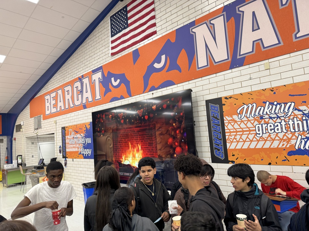 A group of students are gathered around a digital screen showing a cozy fireplace in a school cafeteria. A sign that says "BEARCAT NATION" can be seen on the wall.