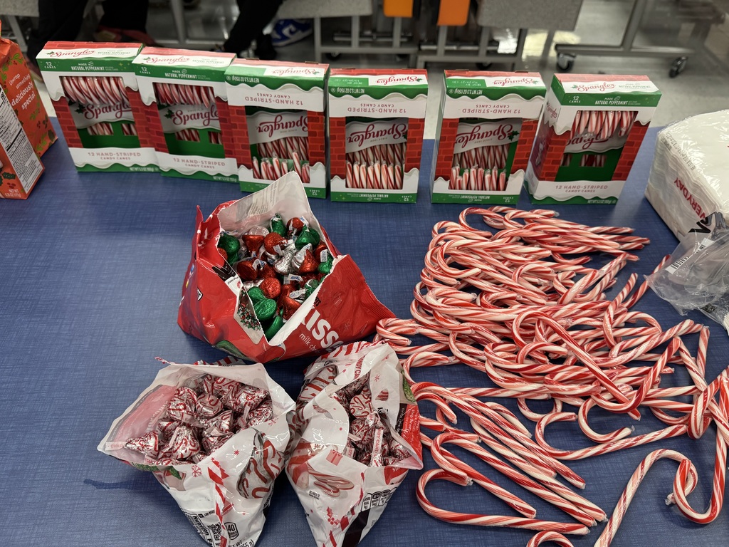 Boxes of candy canes are lined up on a table, with loose candy canes and open bags of colorful foil-wrapped chocolates scattered in front.