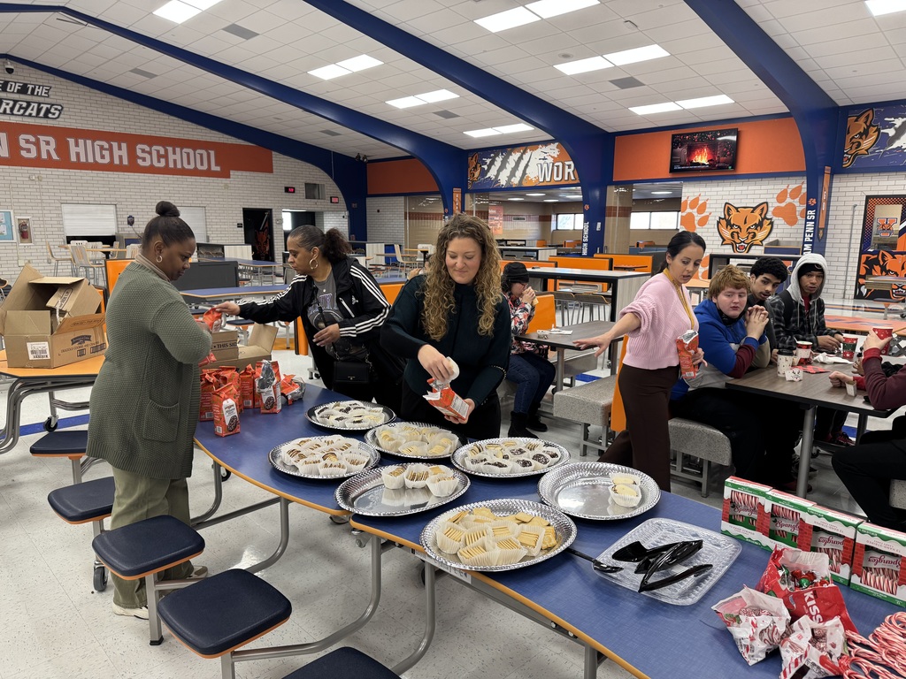 A group of district staff members are setting up a table with assorted pastries and snacks in a school cafeteria. A group of students can be seen sitting nearby at a table having a discussion with a district staff member.