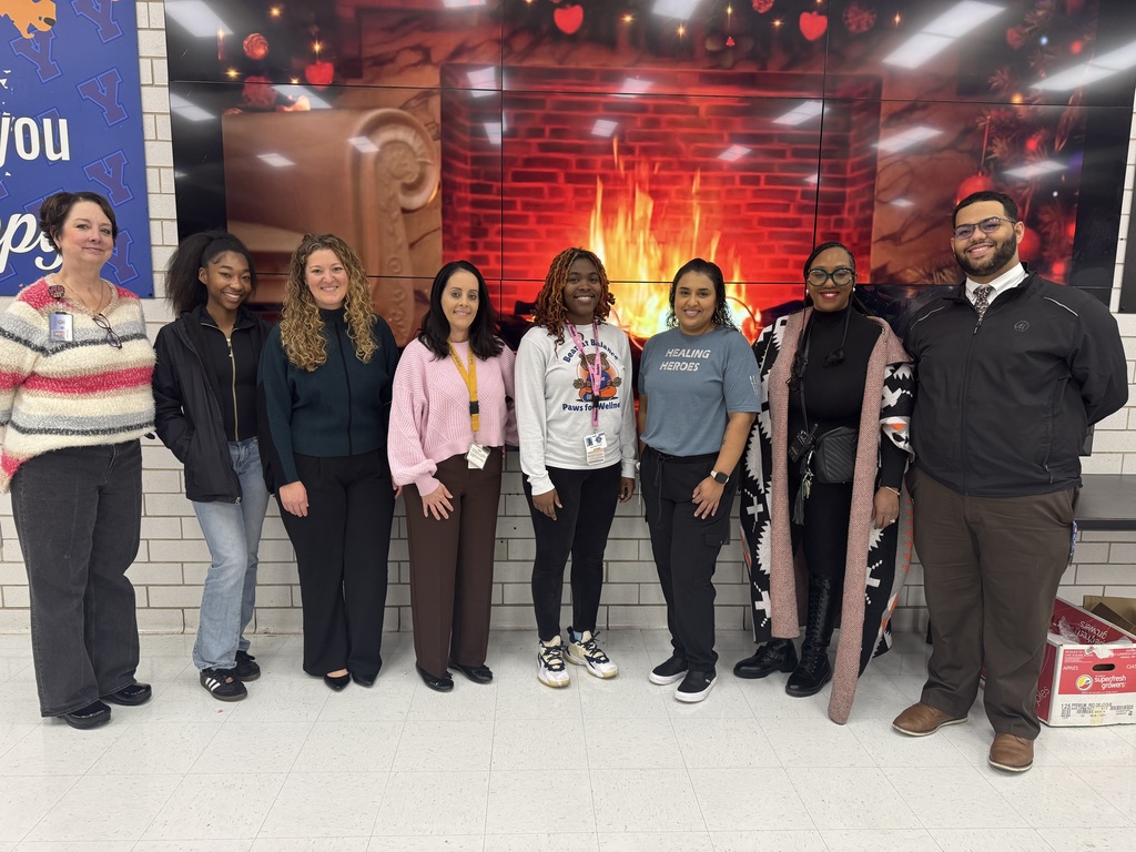 A group of eight district staff members and a student are standing in front of a digital screen displaying a cozy fireplace.