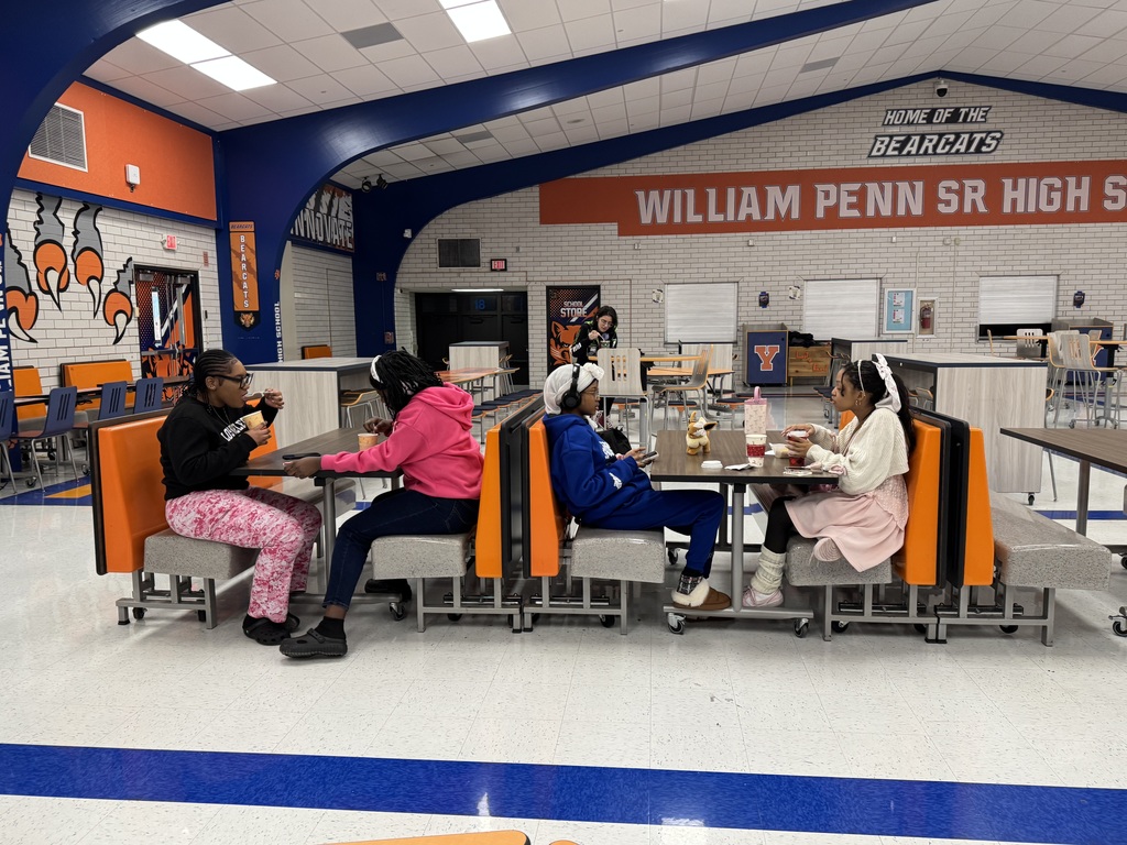 A group of four students are sitting at a cafeteria table in a school cafeteria, eating and talking with one another. There is a sign in the background that reads "Home of the BEARCATS WILLIAM PENN SR HIGH SCHOOL."