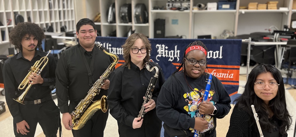 Five band students are standing in a school classroom holding their musical instruments, including a trumpet, saxophone, clarinet, and flute, smiling in a music room.
