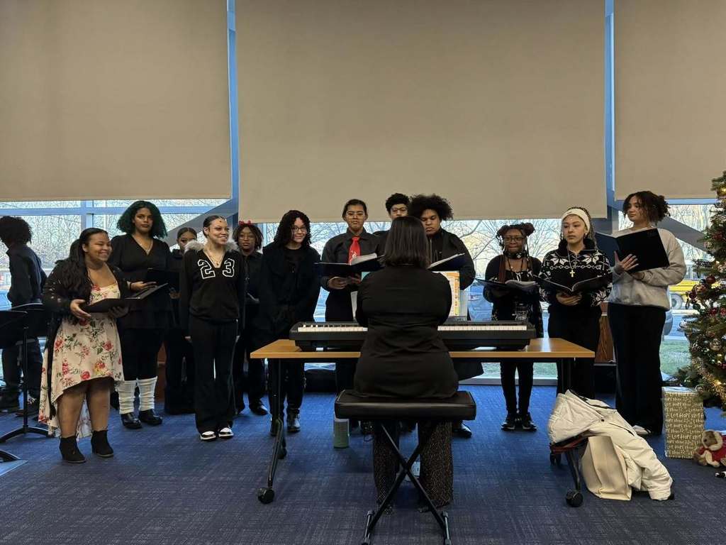 A choir group is standing in a semicircle behind a seated conductor playing a keyboard. They are holding songbooks and performing in a conference room. A decorated Christmas tree is visible to the side, adding a festive touch.