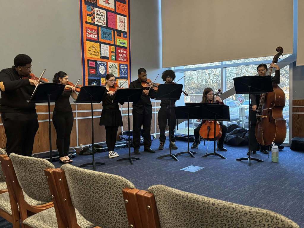 A small ensemble is performing in a conference room with large windows. The group includes violinists, a cellist, and a double bassist. A colorful quilt is hanging on the wall behind them.