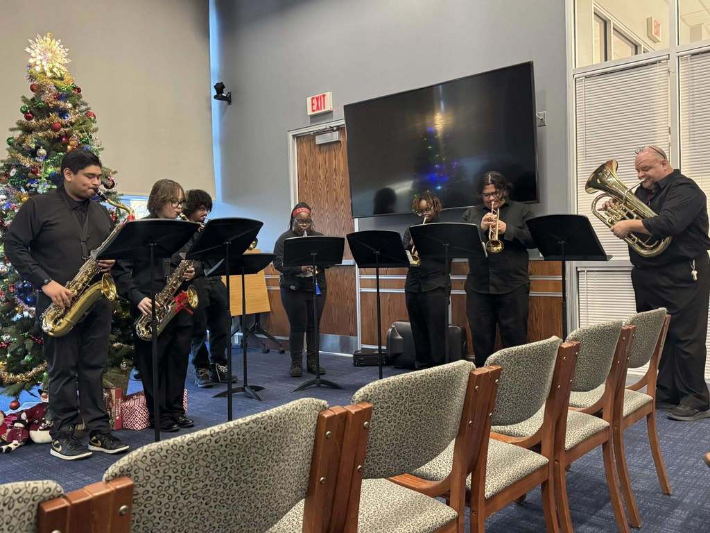 A group of musicians are playing brass and woodwind instruments beside a decorated Christmas tree in a conference room.