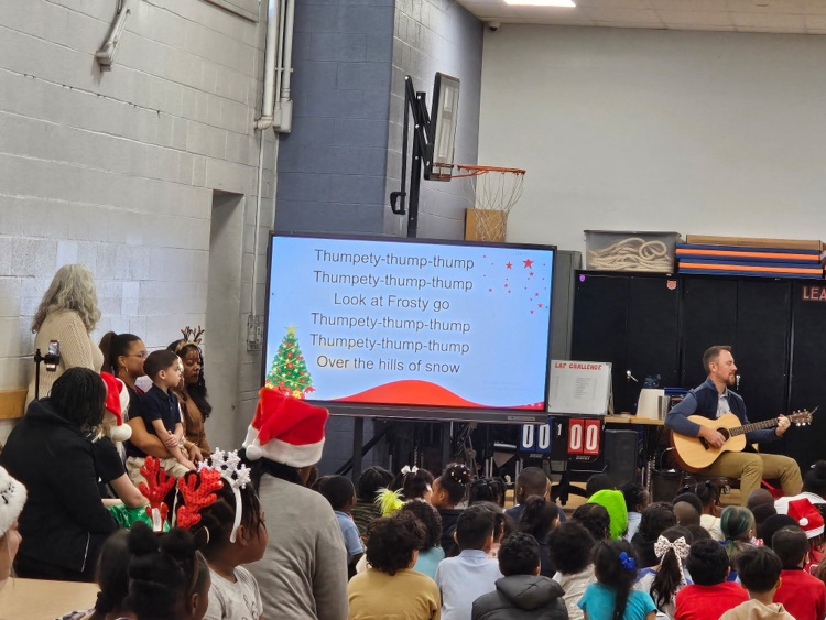 A man is playing a guitar beside a large screen displaying holiday song lyrics. Children in festive attire, including Santa hats and reindeer antlers, are watching eagerly in a school gym.