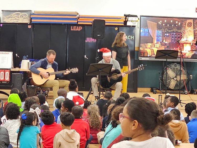 Two musicians are playing acoustic guitars, one wearing a Santa hat, while a woman is standing nearby. Children are sitting on a school gym floor, watching a festive performance.