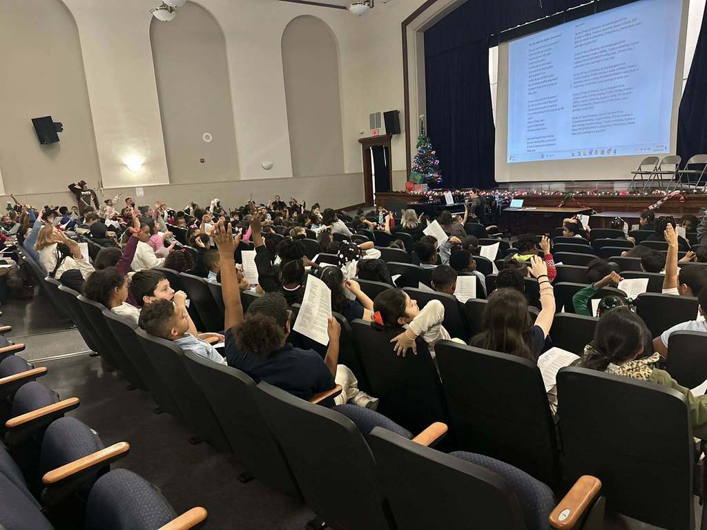 A school auditorium filled with students seated in rows, many raising their hands enthusiastically. A large screen is displaying a text with lyrics to a Christmas song, and a decorated Christmas tree is on stage. A district staff member can be seen sitting at a piano talking into a microphone. 