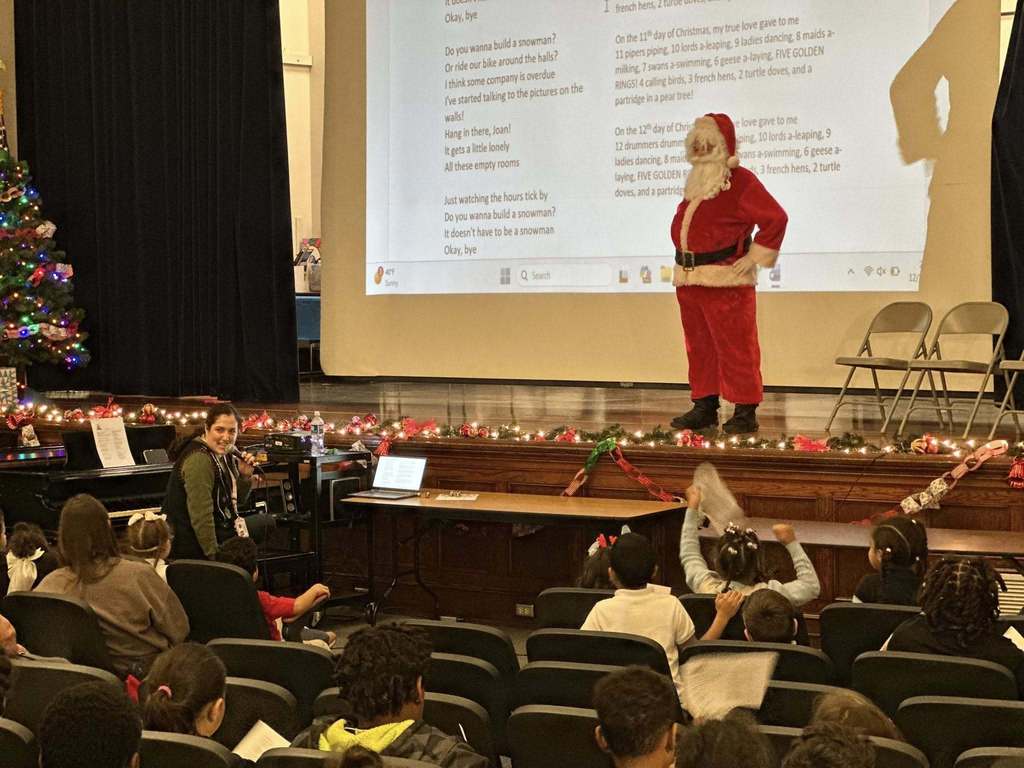 Santa Claus is standing on a decorated stage in a school auditorium, with song lyrics projected behind him. A district staff member can be seen sitting at a piano talking into a microphone. A decorated Christmas tree and festive lights can be seen. A group of students are seated facing the stage and a Christmas tree can be seen nearby. 