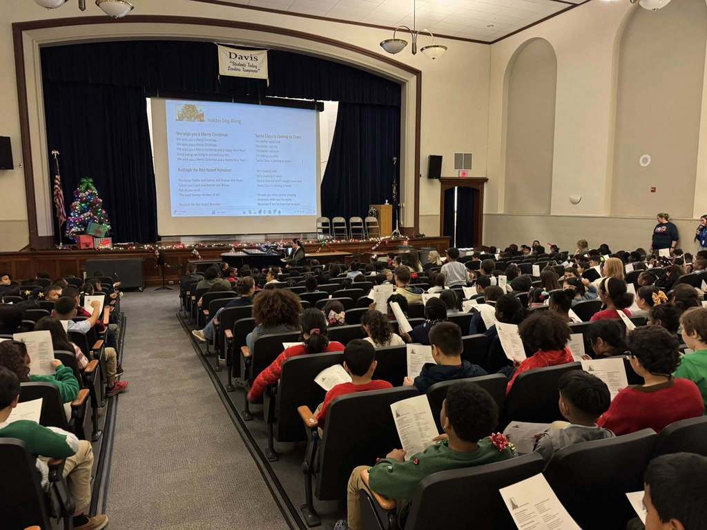 A large group of students in festive attire are sitting in an school auditorium facing a stage and holding papers in their hands. A district staff member can be seen playing on the piano in front of the students. A decorated Christmas tree and festive lights can be seen. A Christmas tree and holiday decorations can be seen.