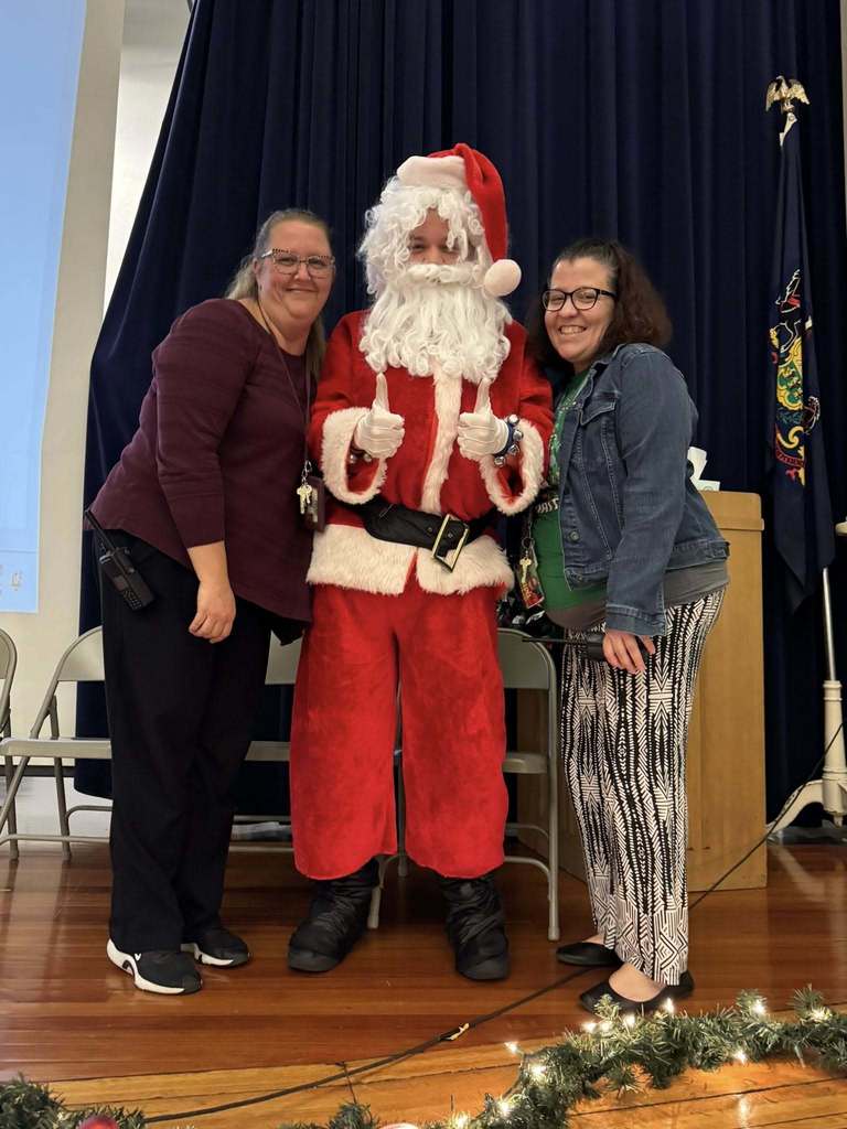 Three district staff members are standing on a stage in a school auditorium. The middle person is dressed as Santa Claus, holding both of their thumbs up. 