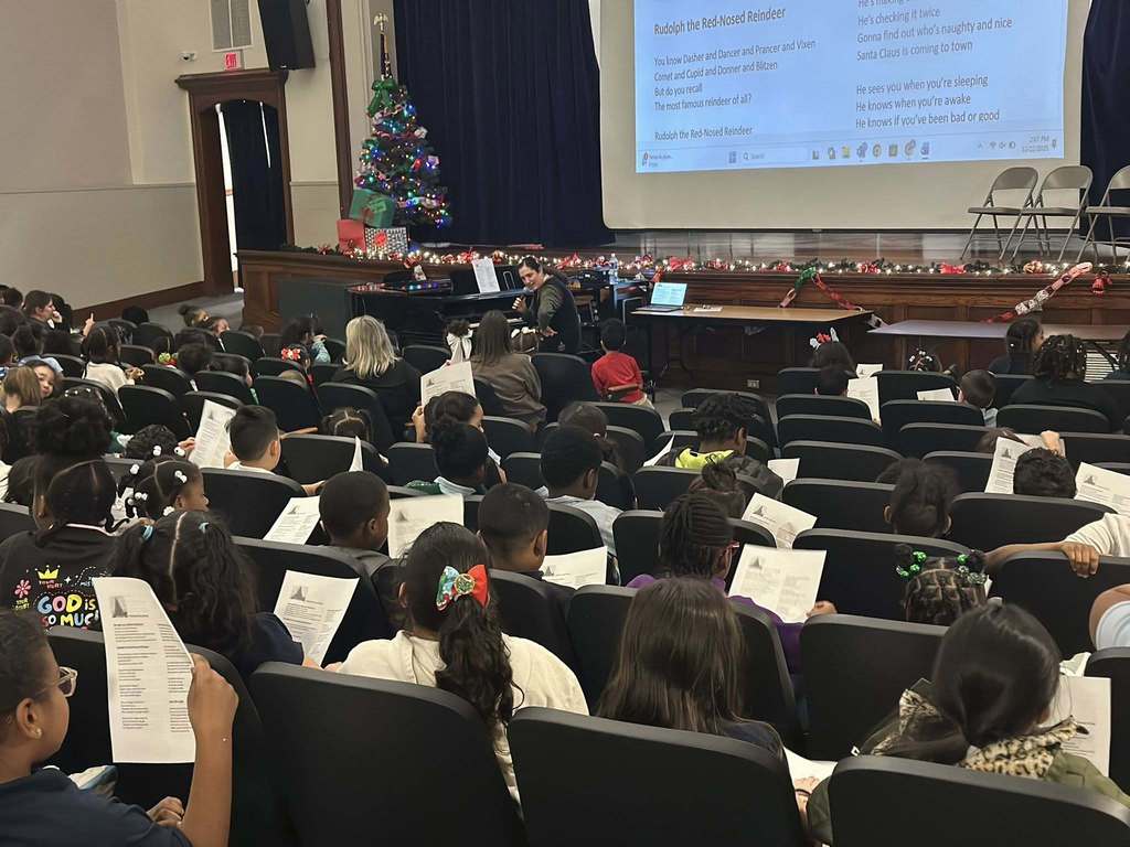 A group of children are sitting in a school auditorium singing along to "Rudolph the Red-Nosed Reindeer." A district staff member can be seen sitting at a piano talking into a microphone. A decorated Christmas tree and festive lights can be seen. 