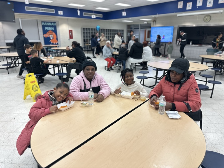 Two women and two young girls are sitting at a table with food and drinks on it in a school cafeteria. Other people can be seen in the background.