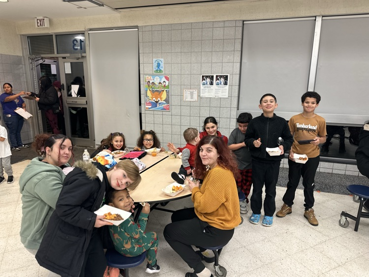 A group of adults and children are standing and sitting at a table with food and drinks on it in a school cafeteria. Other people can be seen in the background.