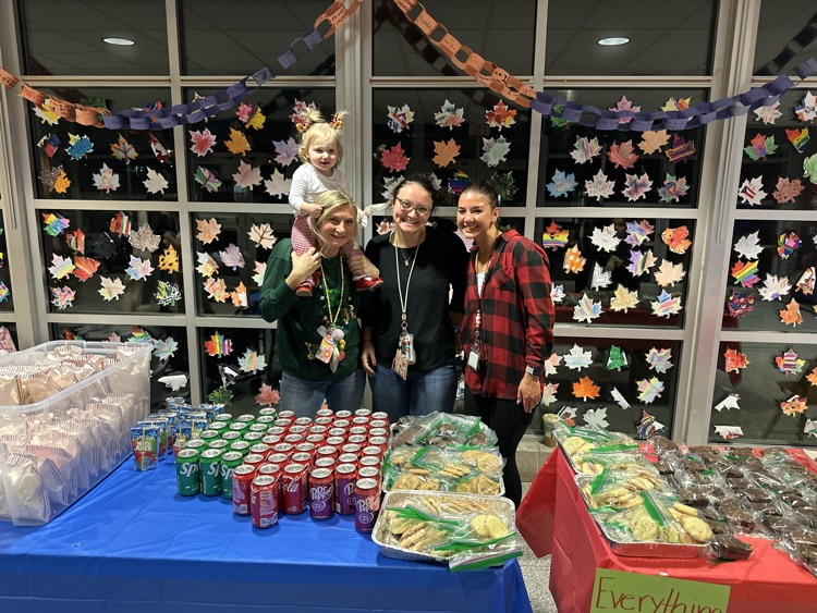 Three district staff members and a young girl are standing behind a table with desserts, canned drinks, and other snacks on it. The young girl is sitting on the shoulders of one of the district staff members.