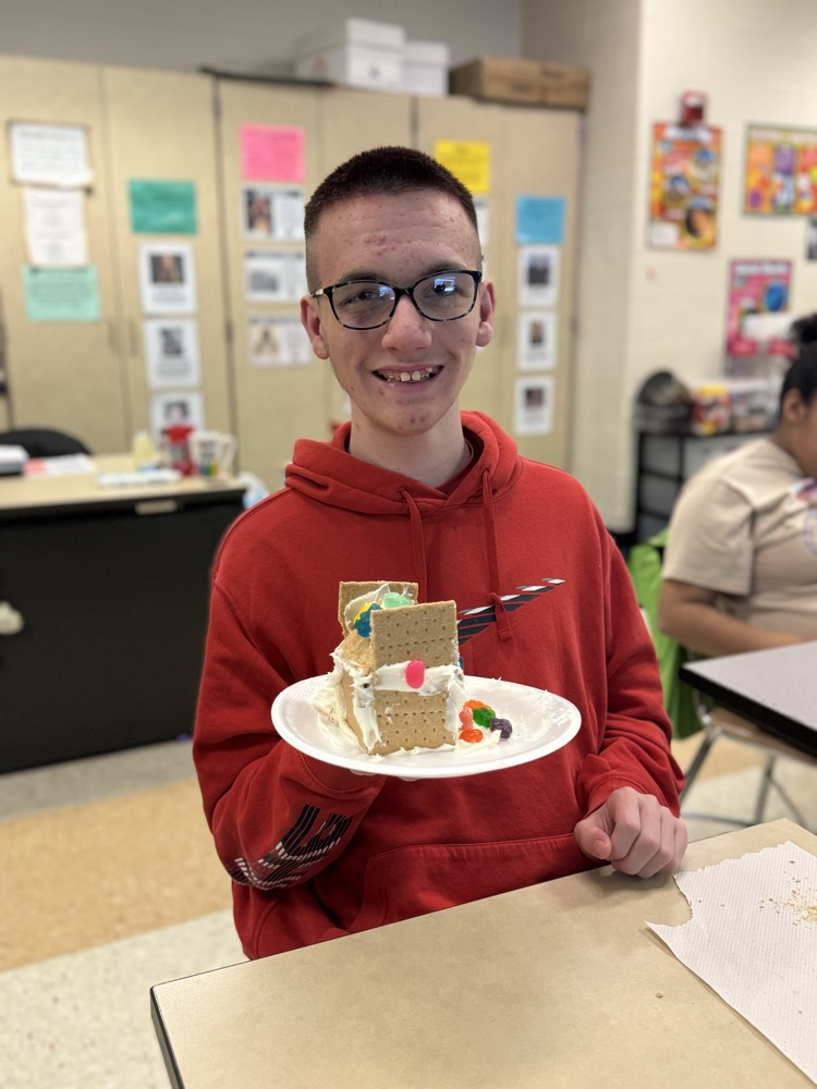 A student is sitting at a desk in a school classroom, holding up a gingerbread house. Other people can be seen in the background.