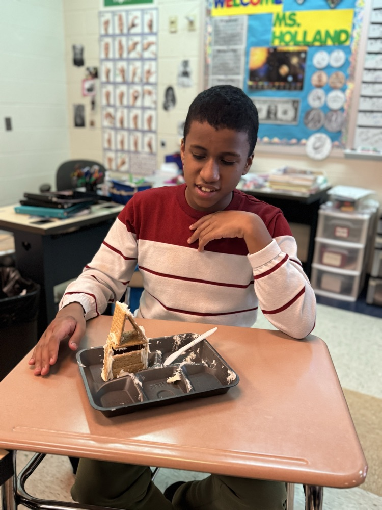 A student is sitting at a desk in a school classroom, looking down at a gingerbread house.