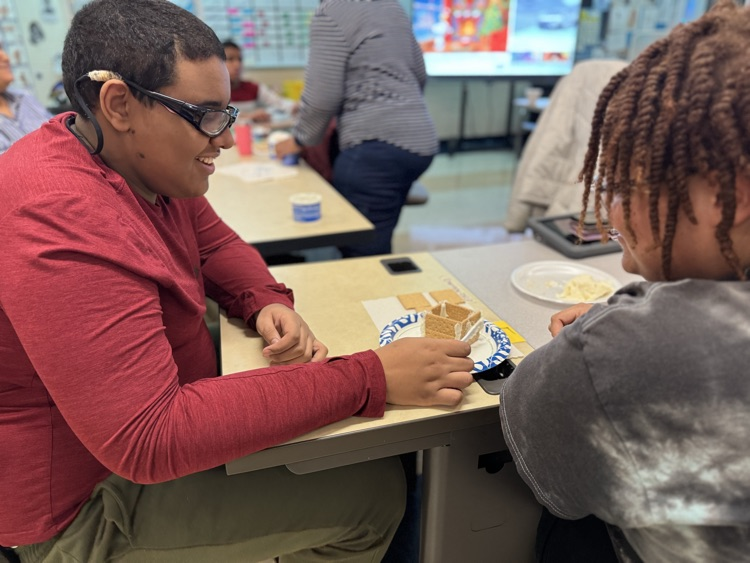 A district staff member and a student are decorating a gingerbread house in a school classroom. Other people can be seen in the background.