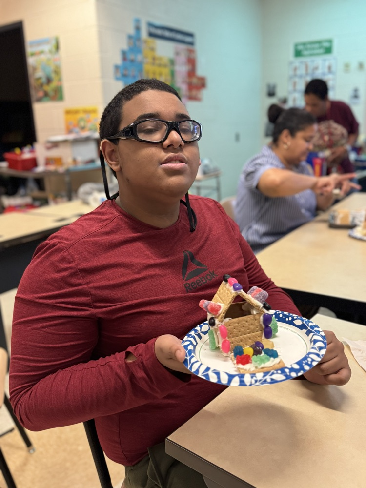 A student is sitting at a desk in a school classroom, holding up a gingerbread house. Other people can be seen in the background.