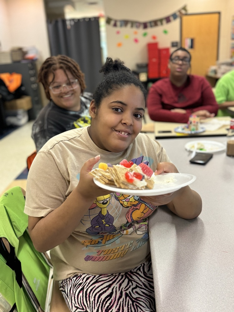 A student is sitting at a desk in a school classroom, holding up a gingerbread house. Other people can be seen in the background.