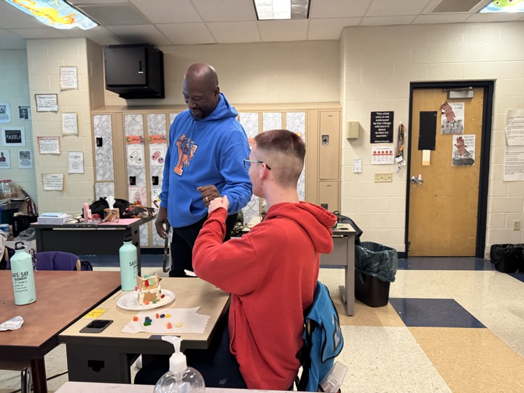 A district staff member is giving a fist bump to a student who is sitting at a desk in a school classroom.