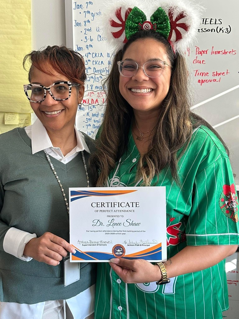 Jackson PreK-8 Principal, Ms. Karen Smallwood, is standing next to a district staff member, both of them are holding a certificate of perfect attendance. The district staff member is wearing a holiday headband and festive shirt. A whiteboard can be seen in the background.
