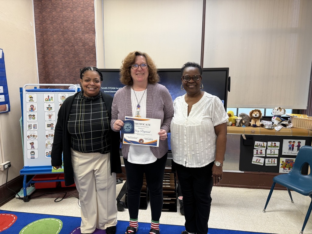 Superintendent of Schools, Dr. Berry-Brown, is standing with a district staff member and a community member in a school classroom. The district staff member is holding a certificate and a cup coaster in her hand.