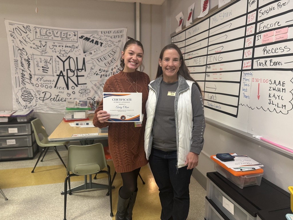 A district staff member is standing next to a community member in a school classroom. The district staff member is holding a certificate in her hand.