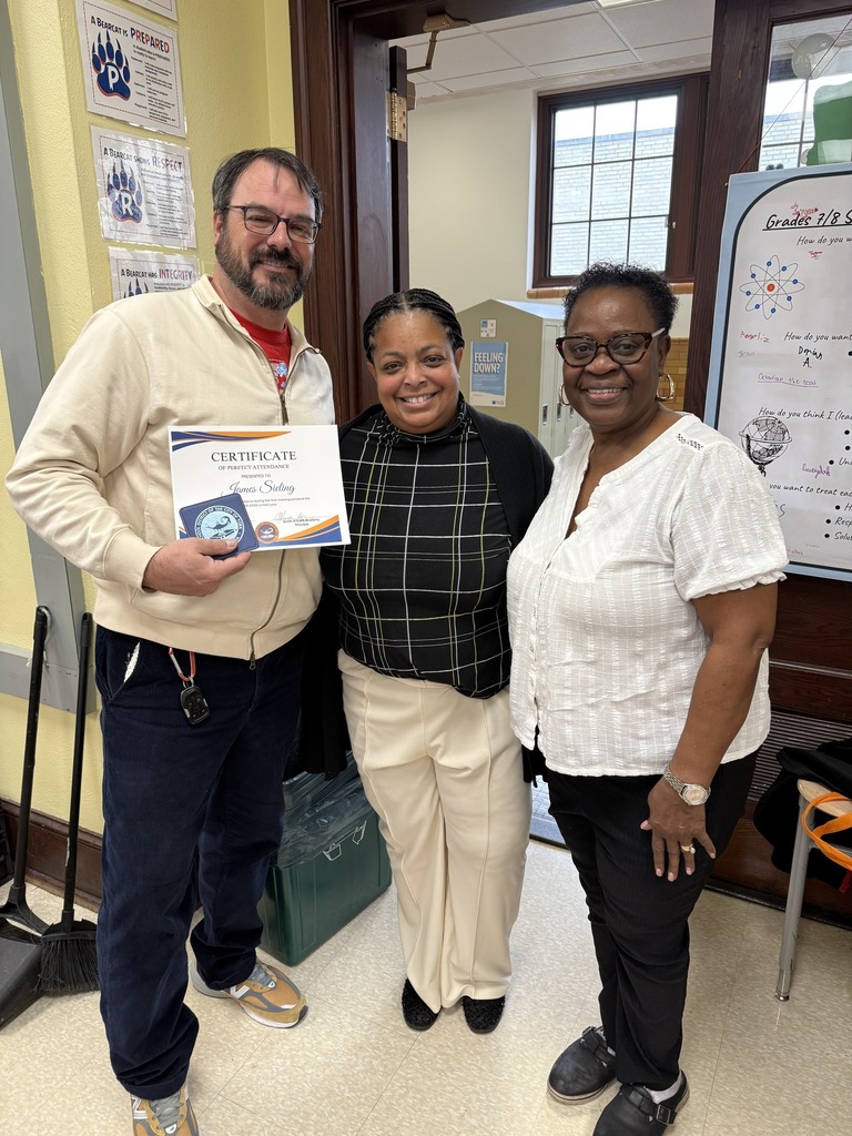 Superintendent of Schools, Dr. Berry-Brown, is standing with a district staff member and a community member in a school classroom. The district staff member is holding a certificate and a cup coaster in his hand.
