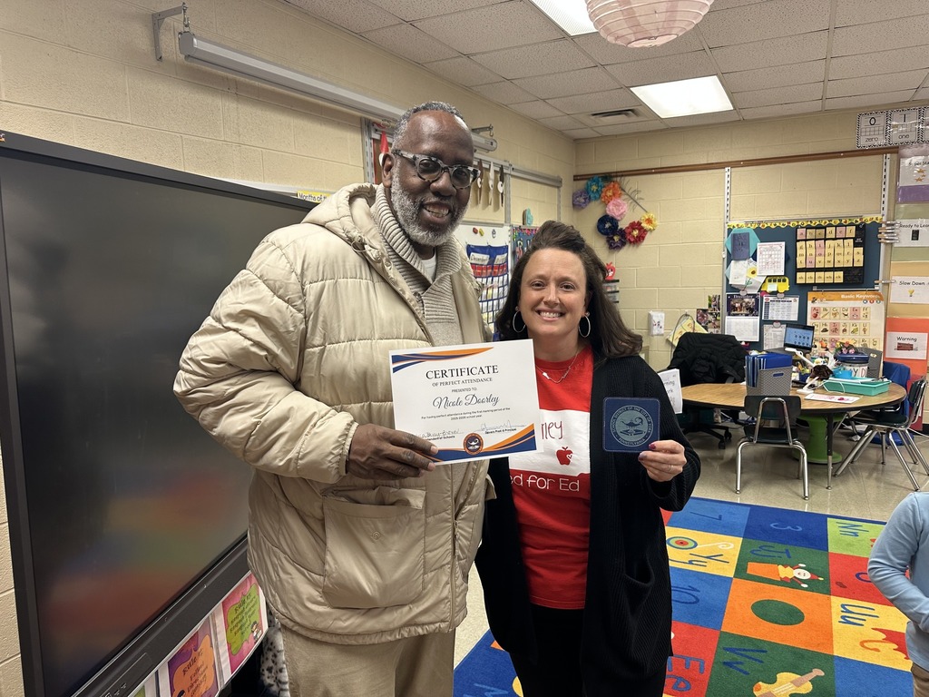 Two district staff members are standing in a classroom. One of the district staff members is holding a certificate, while the other district staff member is holding a cup coaster.
