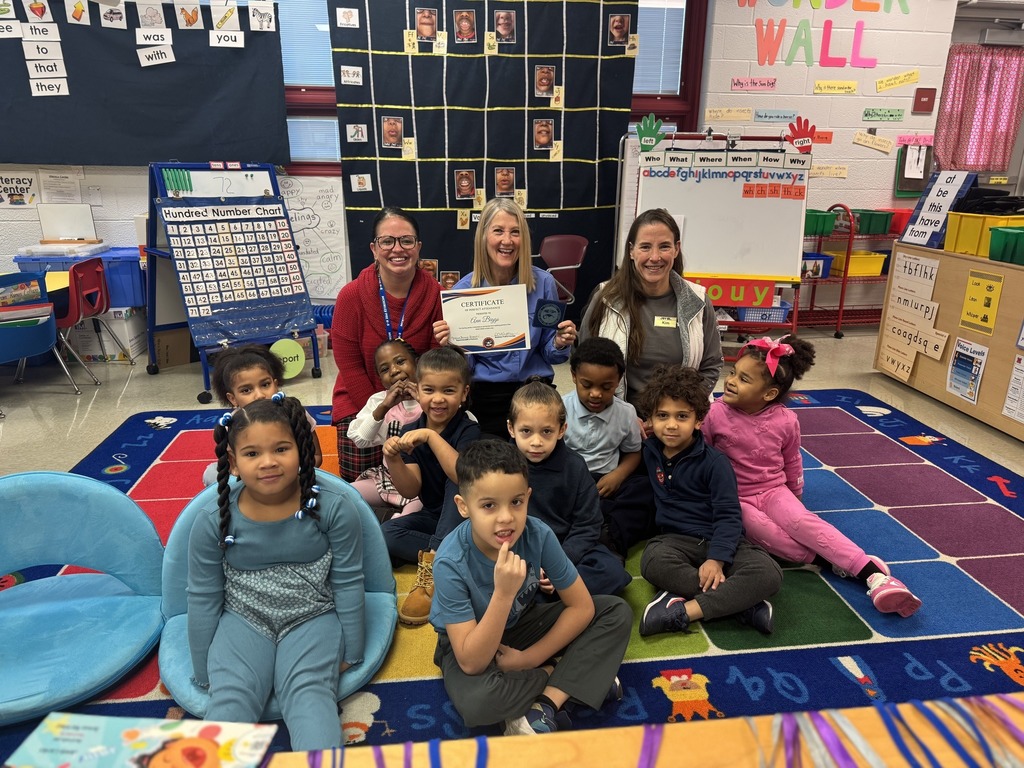 McKinley PreK-8's Principal, Dr. Ashley White, along with a group of students, a district staff member, and a community member are sitting on a colorful carpet in a school classroom. The district staff member is holding a certificate in her hand.