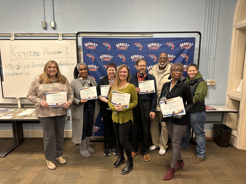 A group of eight district staff members are holding certificates in front of a "Bearcat Cyber" backdrop. A whiteboard reads “Keystone Algebra I.”