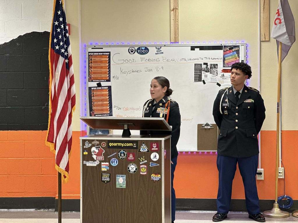 Two students in military uniforms are standing at a podium in a school classroom. An American flag is to their left, and a whiteboard filled with notes is behind them.